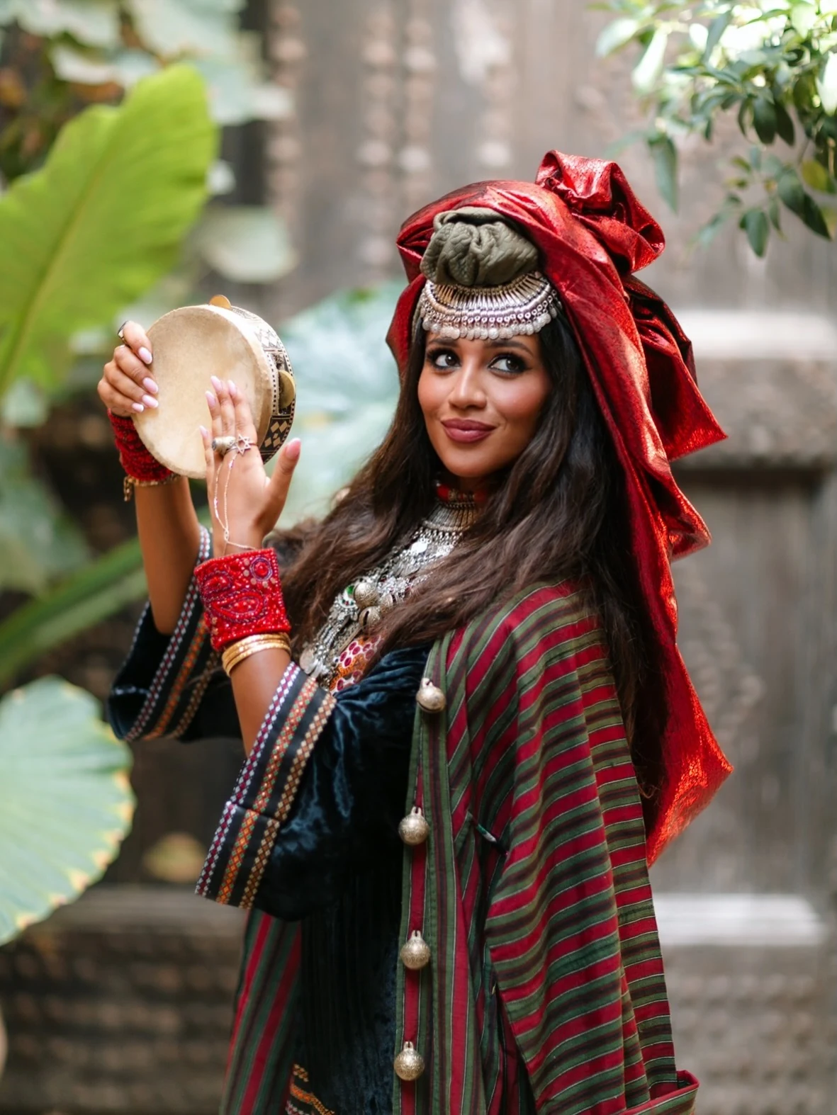 Yzabela holding a frame drum in a leafy courtyard, wearing a red wrap and a green and red striped mantle