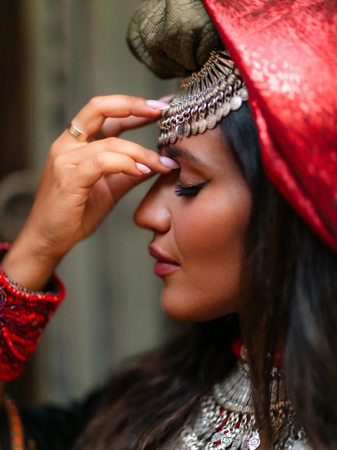 Yzabela in profile, one hand resting beside a silver-colored headpiece and red head covering