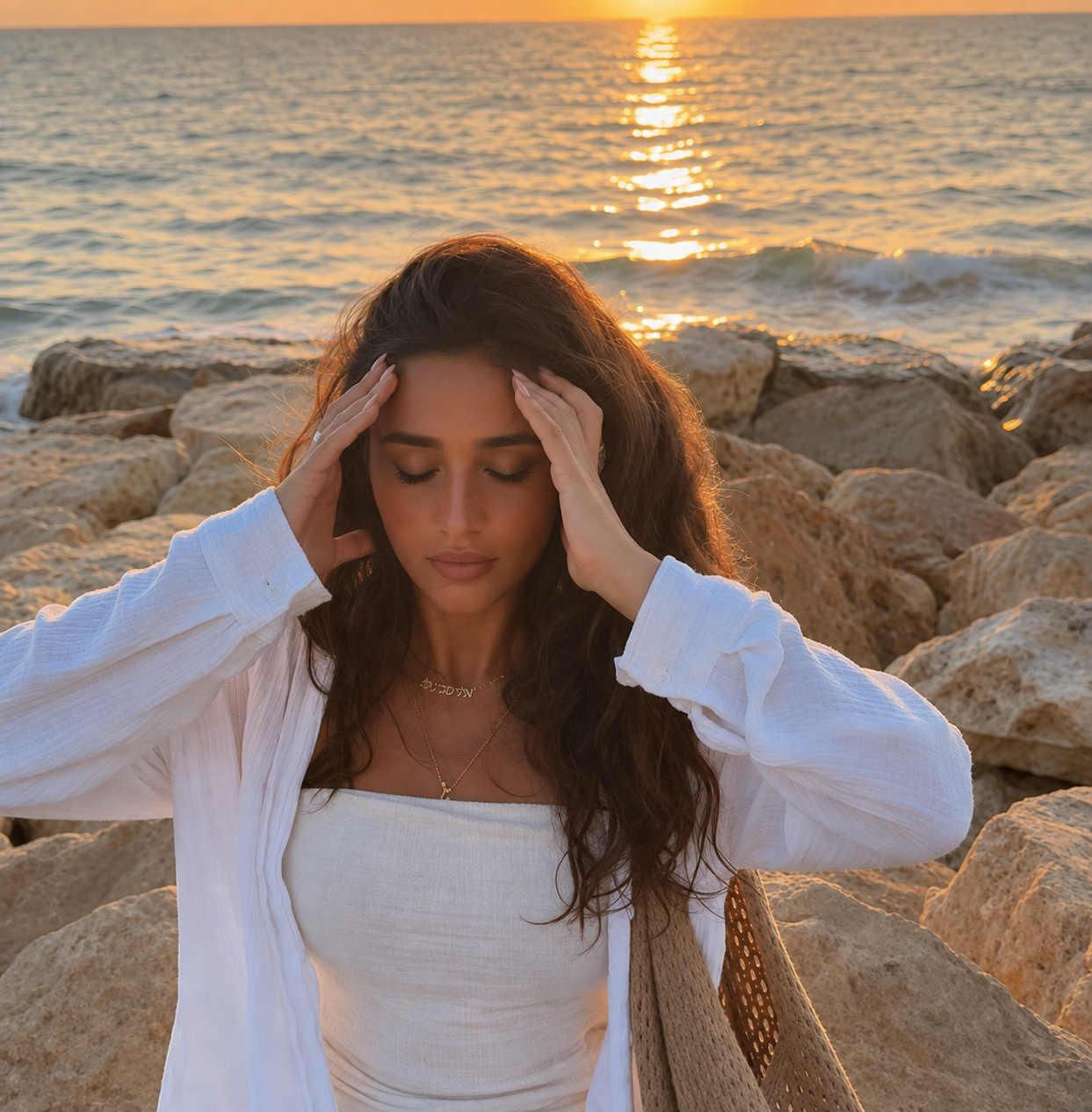 A woman in white seated among shoreline rocks at sunset with her eyes closed and the sea behind her
